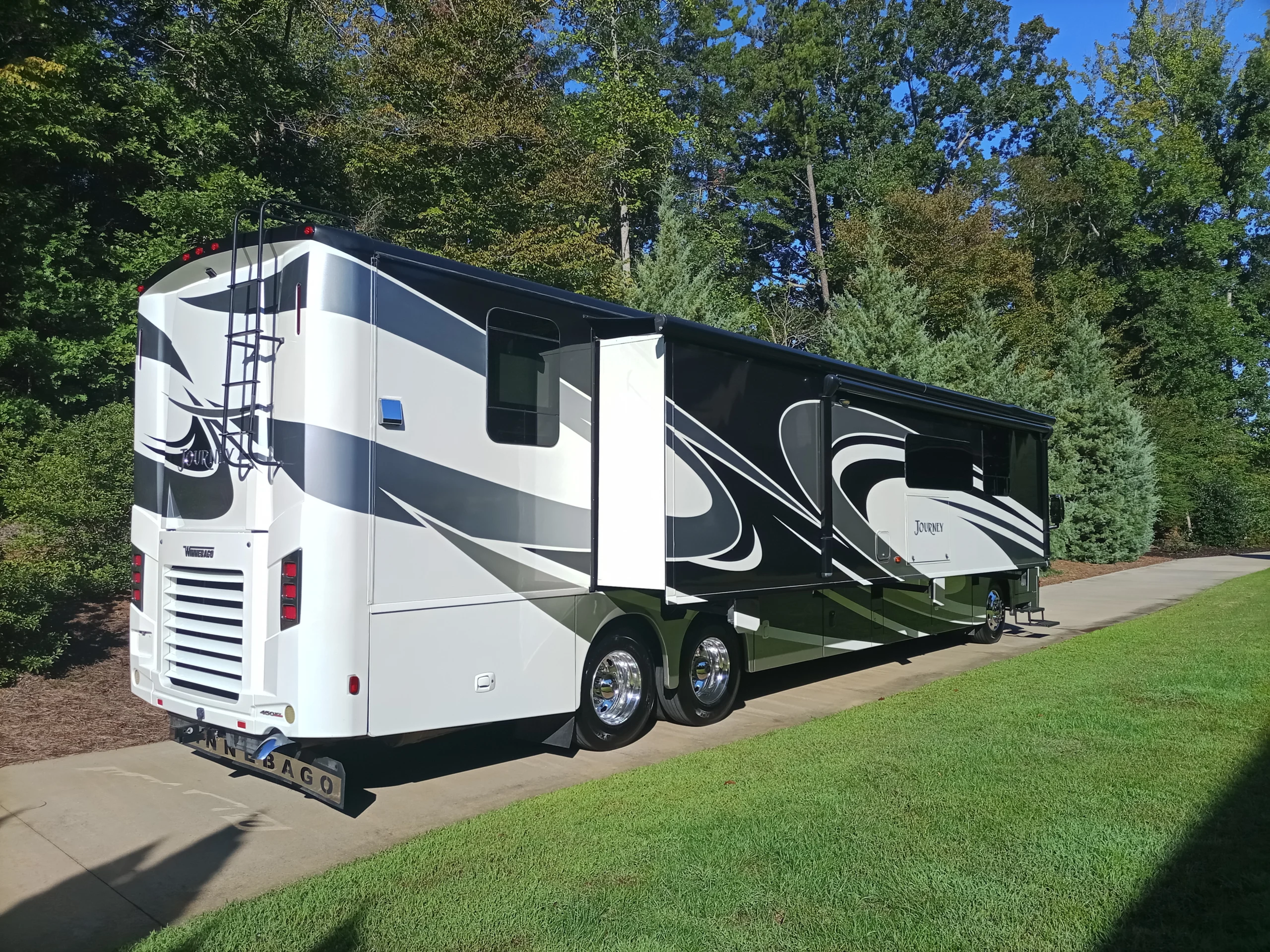 IMG_20260928_094925_638 Luxury motorcoach parked on driveway, surrounded by lush greenery, displaying sleek black and white design patterns.
