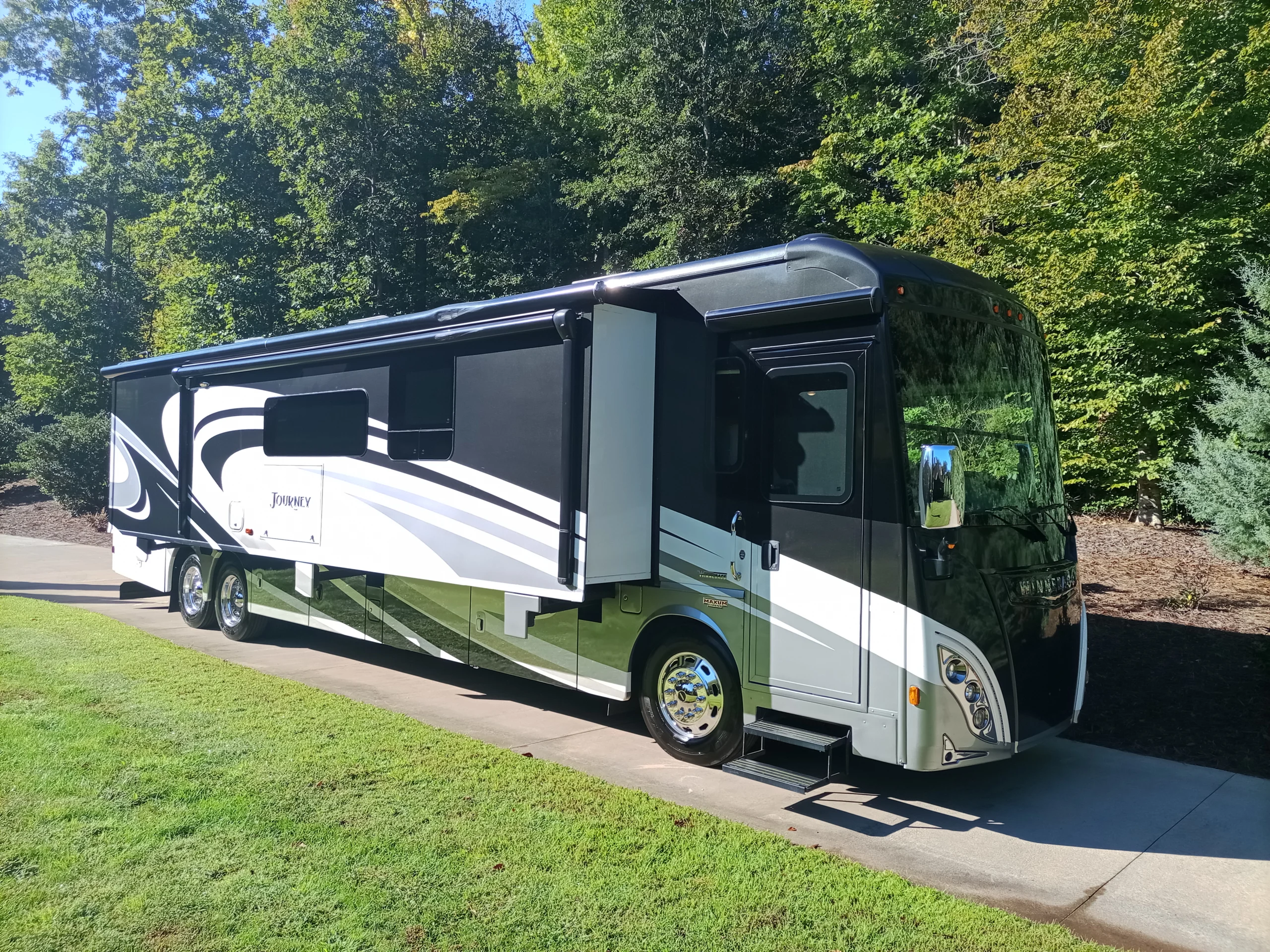 IMG_20260928_094946_568 Luxurious motorcoach parked on a driveway, surrounded by lush green trees, under clear blue skies.