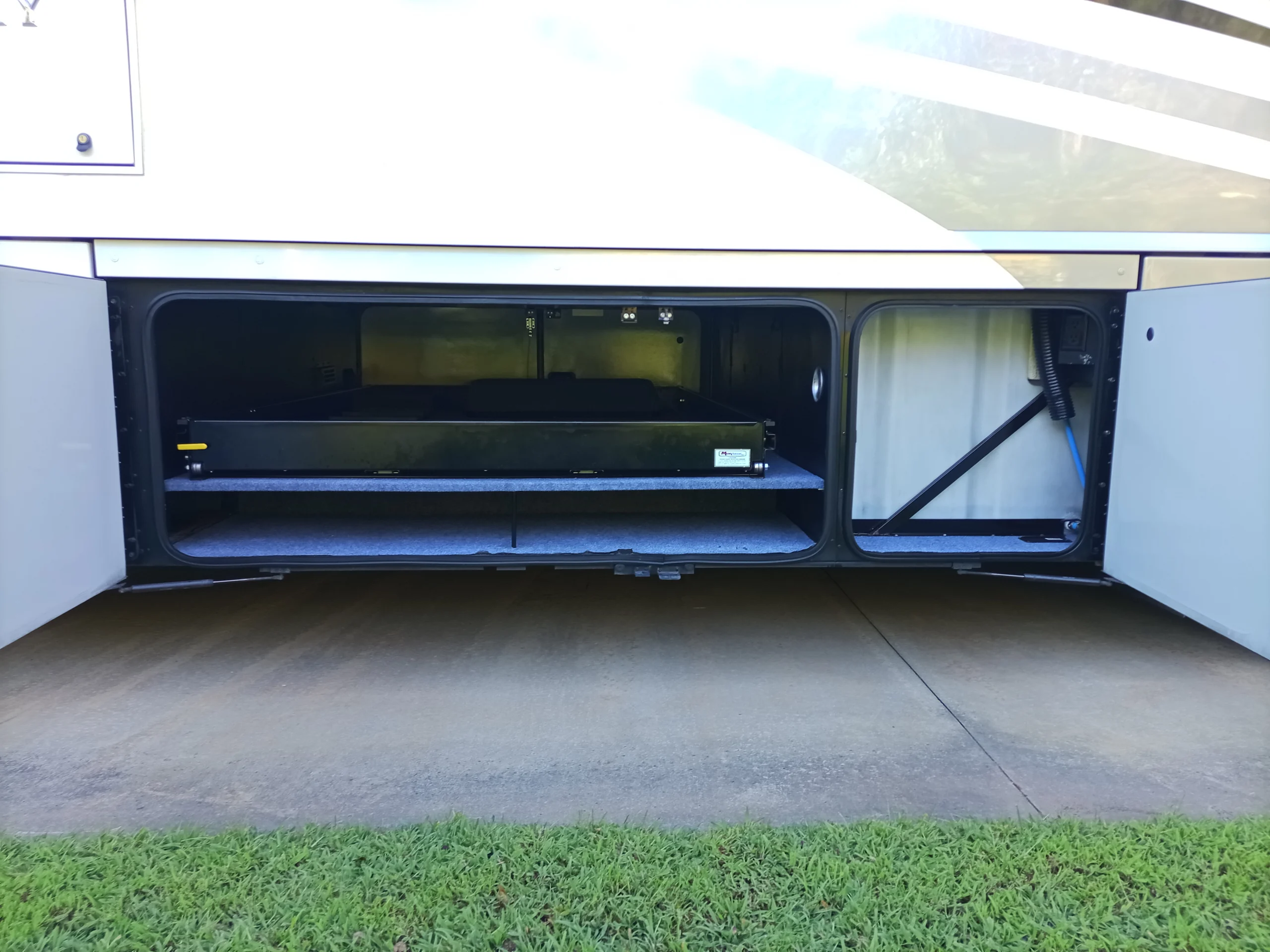 IMG_20260928_160031_382 Open storage compartment of a Motorcoach and RV, revealing spacious interior with organized compartments, over a grassy and paved surface.