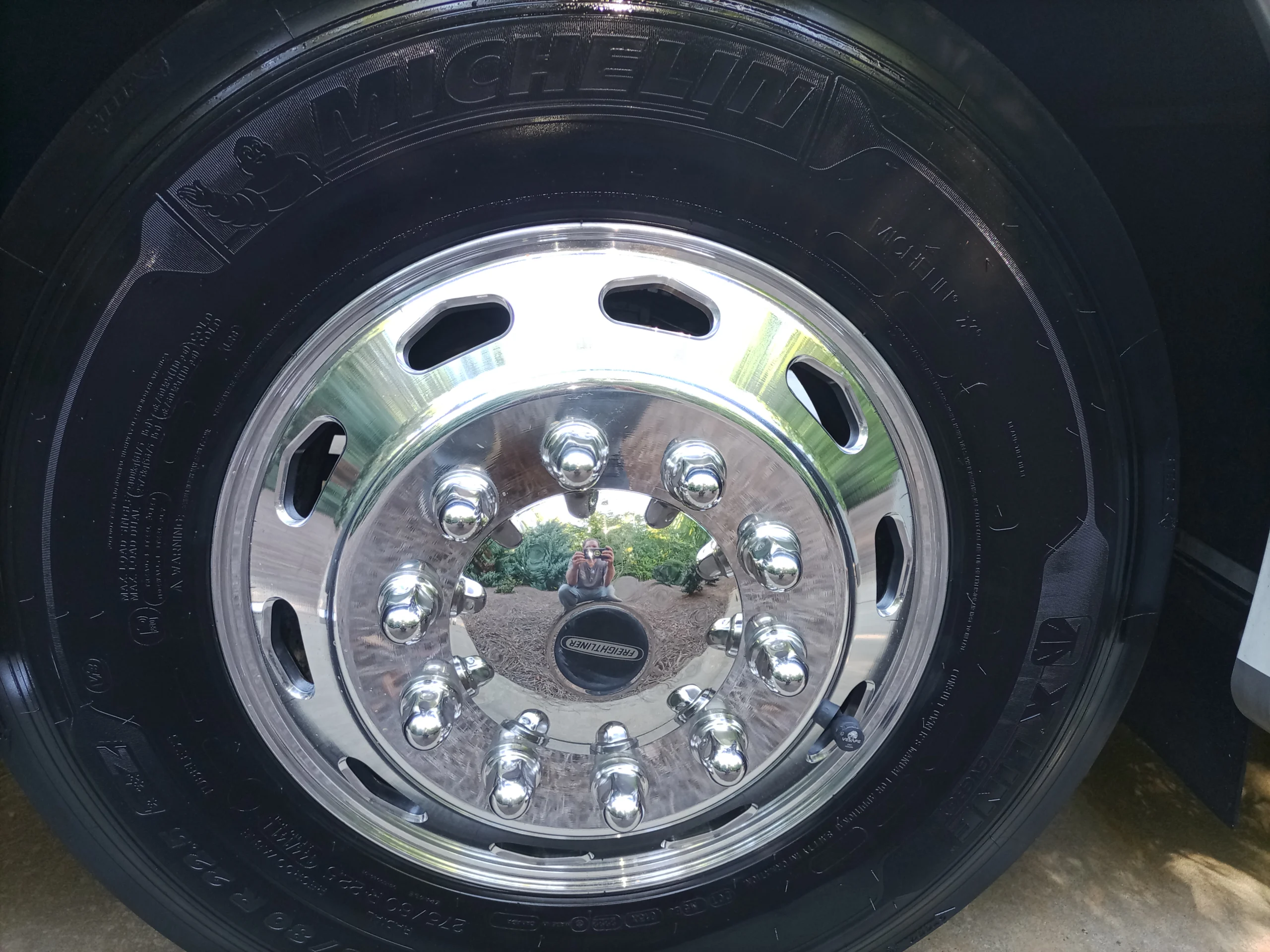 IMG_20260928_160328_054 Shiny chrome wheel of a motorcoach reflecting scenery and a person, Michelin tire with visible branding and bolts.