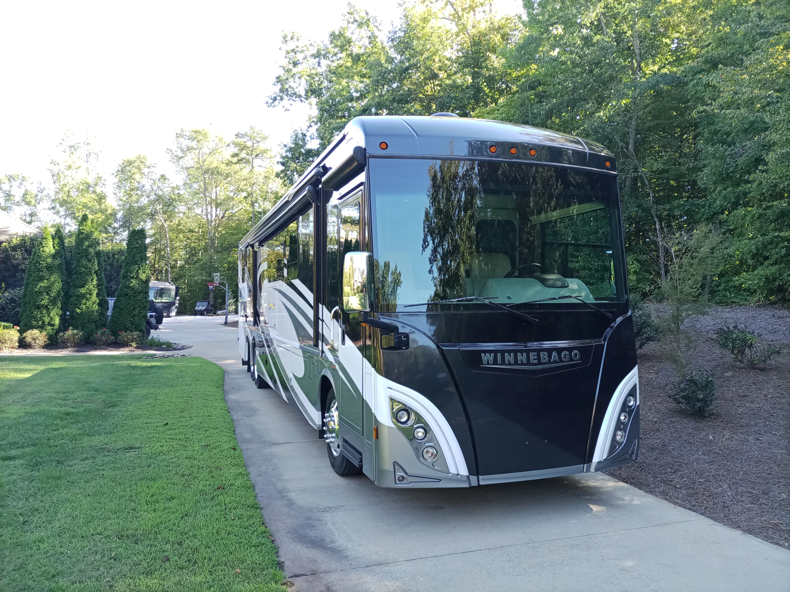 IMG_20260928_160605_477 Winnebago motorcoach parked on a paved driveway surrounded by lush greenery and trees.