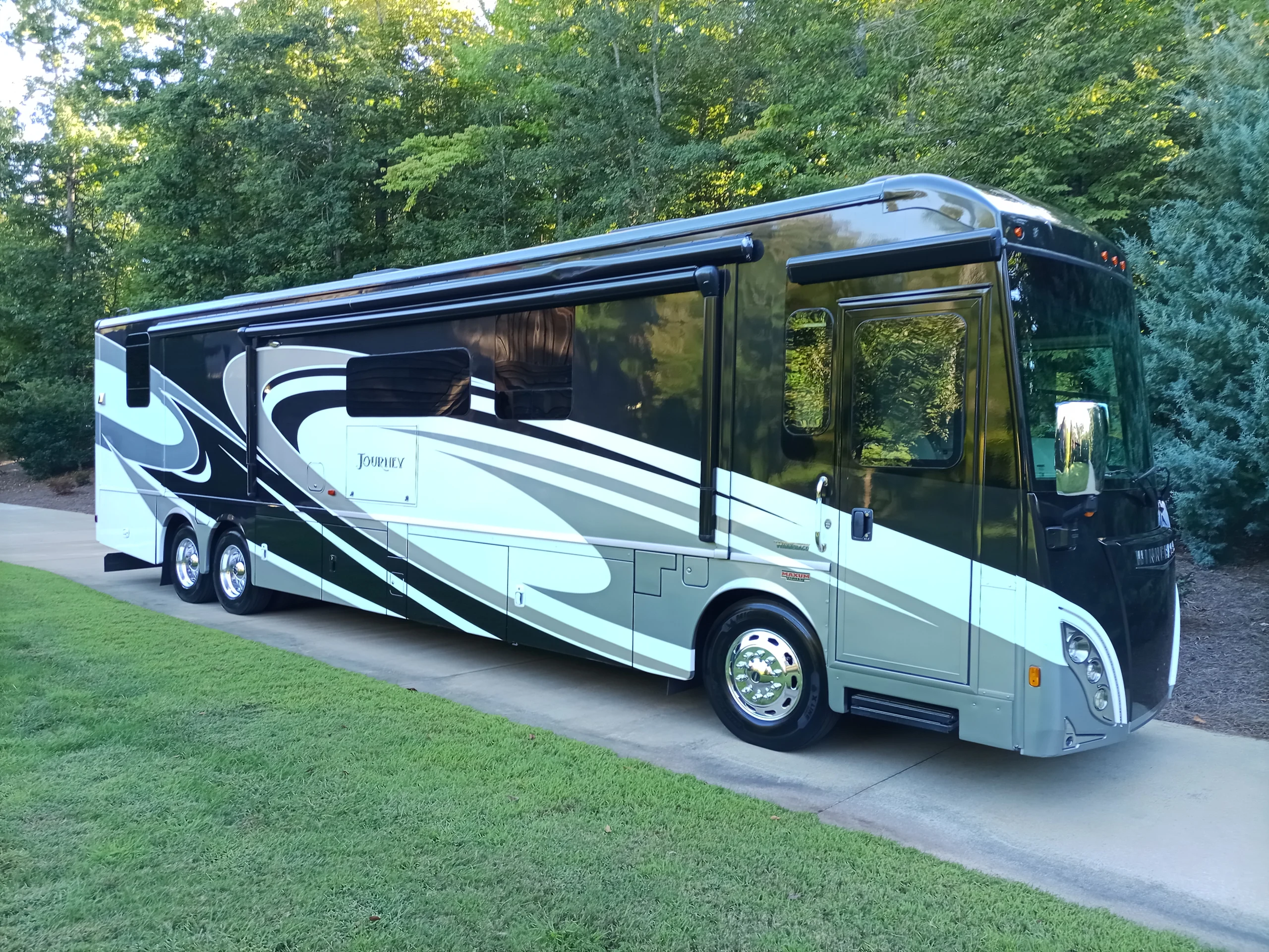 IMG_20260928_170349_280 Luxurious motorcoach parked on a driveway, featuring sleek black and white design, surrounded by lush greenery.