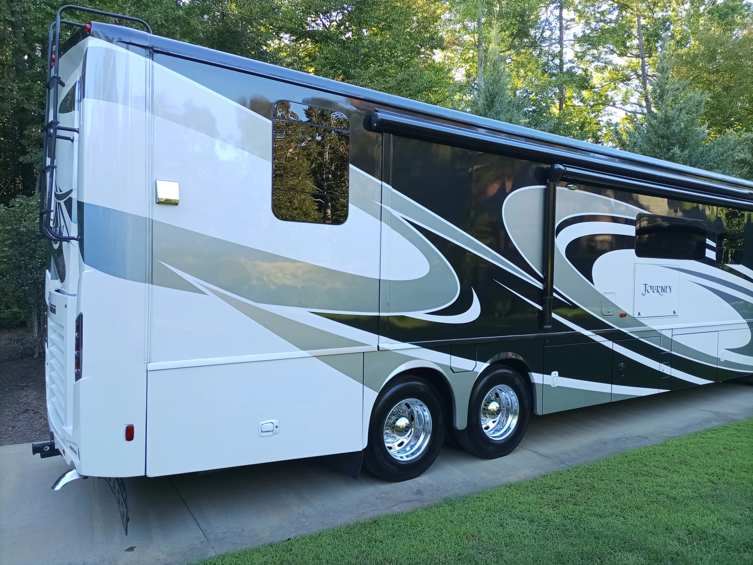 IMG_20260928_170445_559 Motorcoach parked on driveway with sleek swirled design, glossy finish, surrounded by lush green trees and grass.