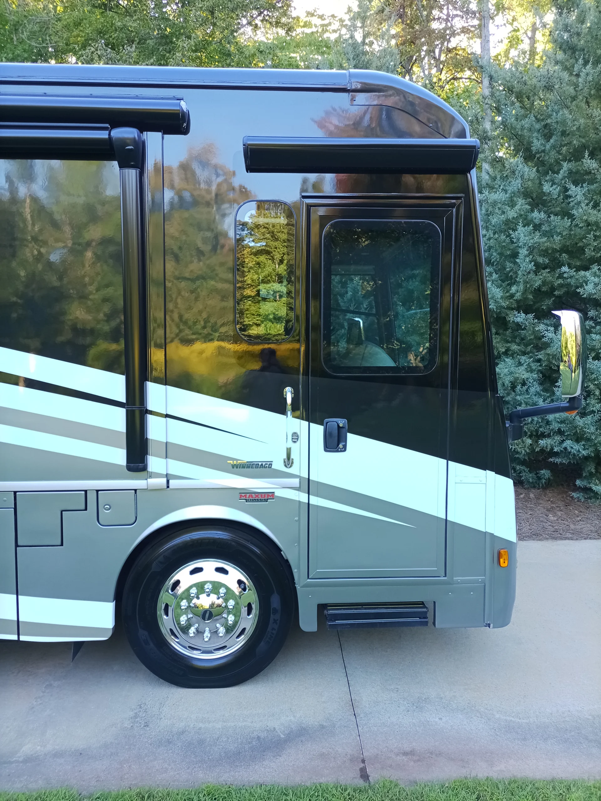 IMG_20260928_170511_366 Front side view of a Motorcoach featuring a sleek design, large mirrored wheel, and retractable awning, set against a lush, green backdrop.
