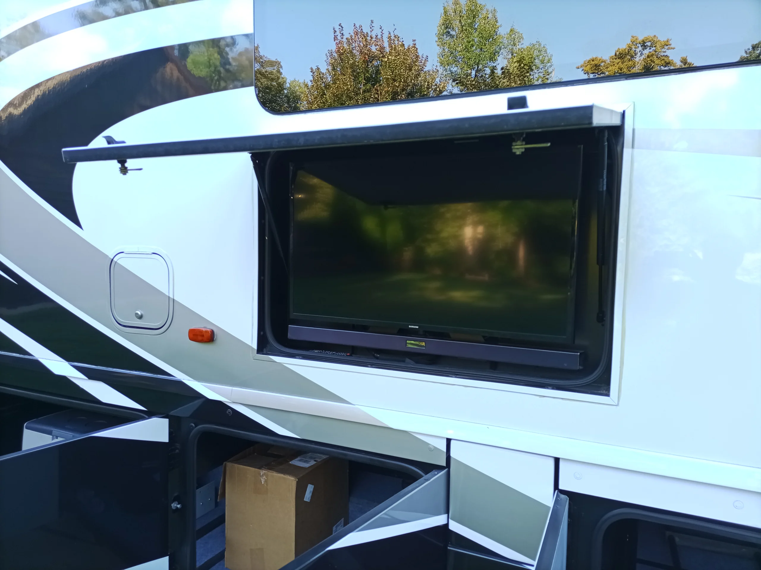 IMG_20260928_170655_168 Outdoor TV in a motorcoach and RV open compartment, with storage below and trees reflecting on the screen.