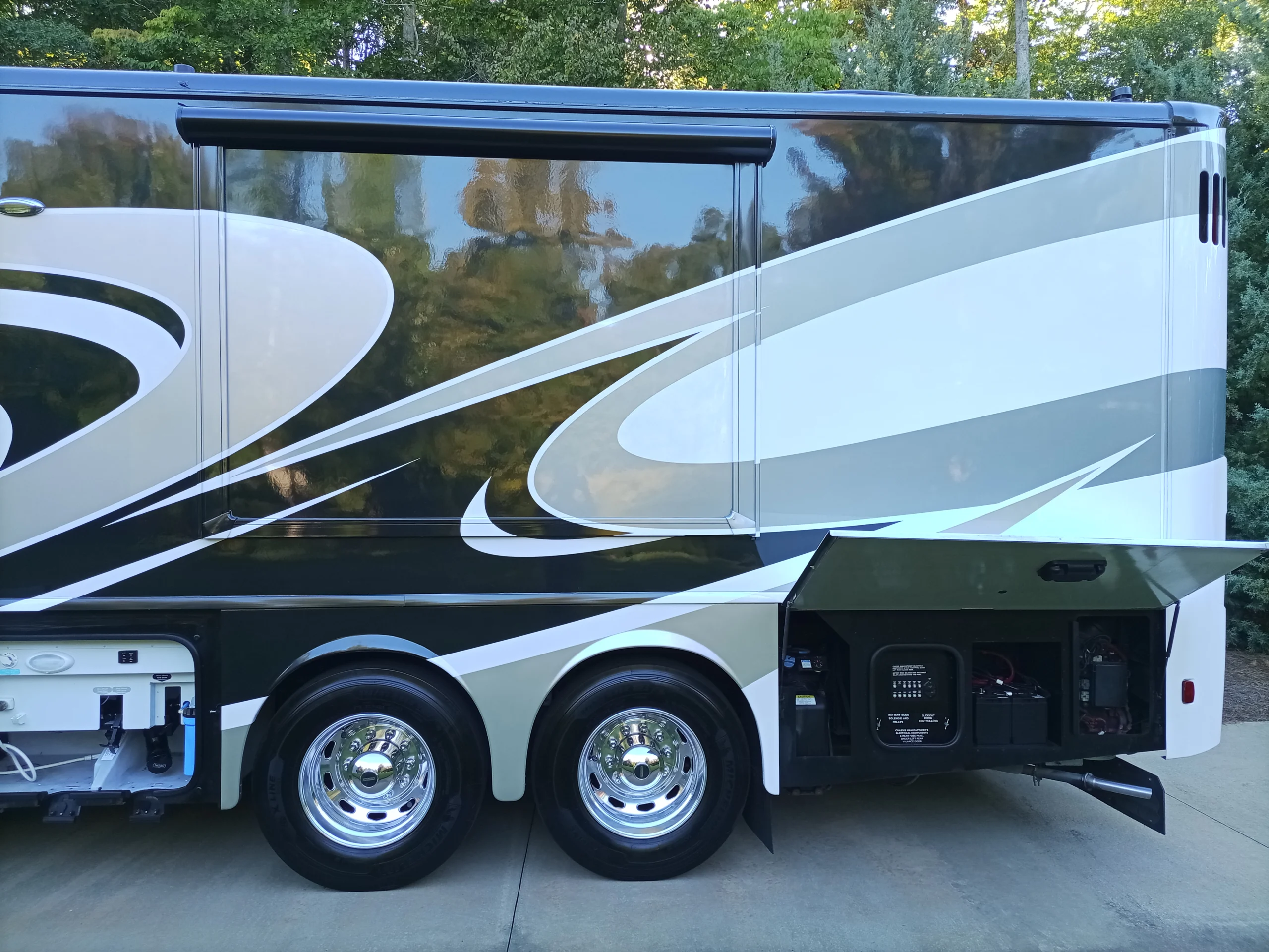 IMG_20260928_171153_970 Side view of a Motorcoach showing polished wheels, external storage compartments, and a decorative swirl pattern, set against greenery.