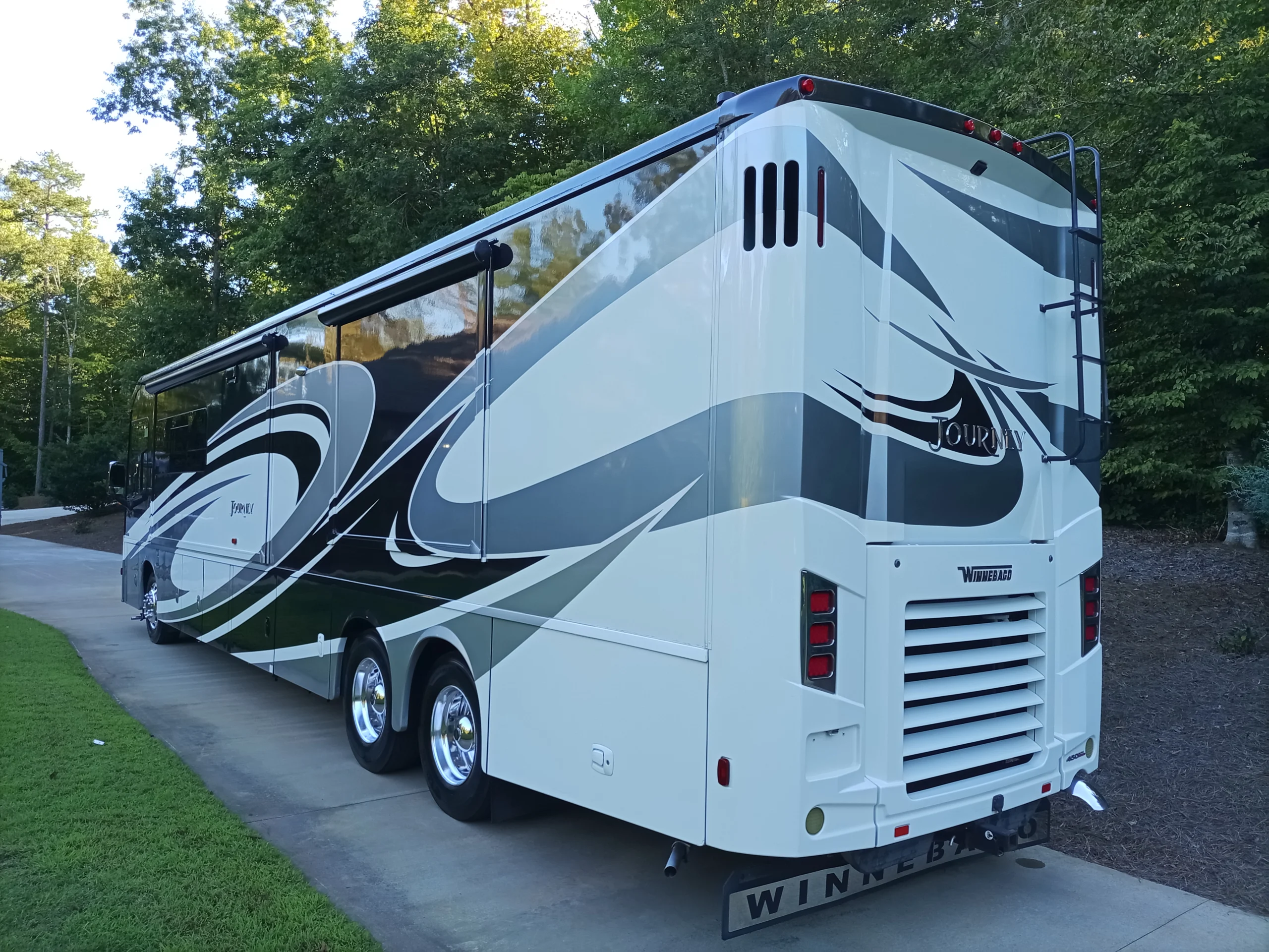 IMG_20260928_171253_355 White and gray motorcoach with curved black accents parked on a driveway surrounded by green trees and grass.